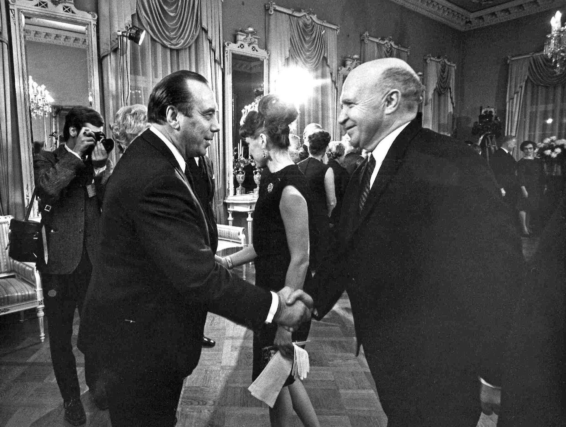 In a decorated room, festively clothed men and women meet, two men in dark suits shake hands in the foreground.