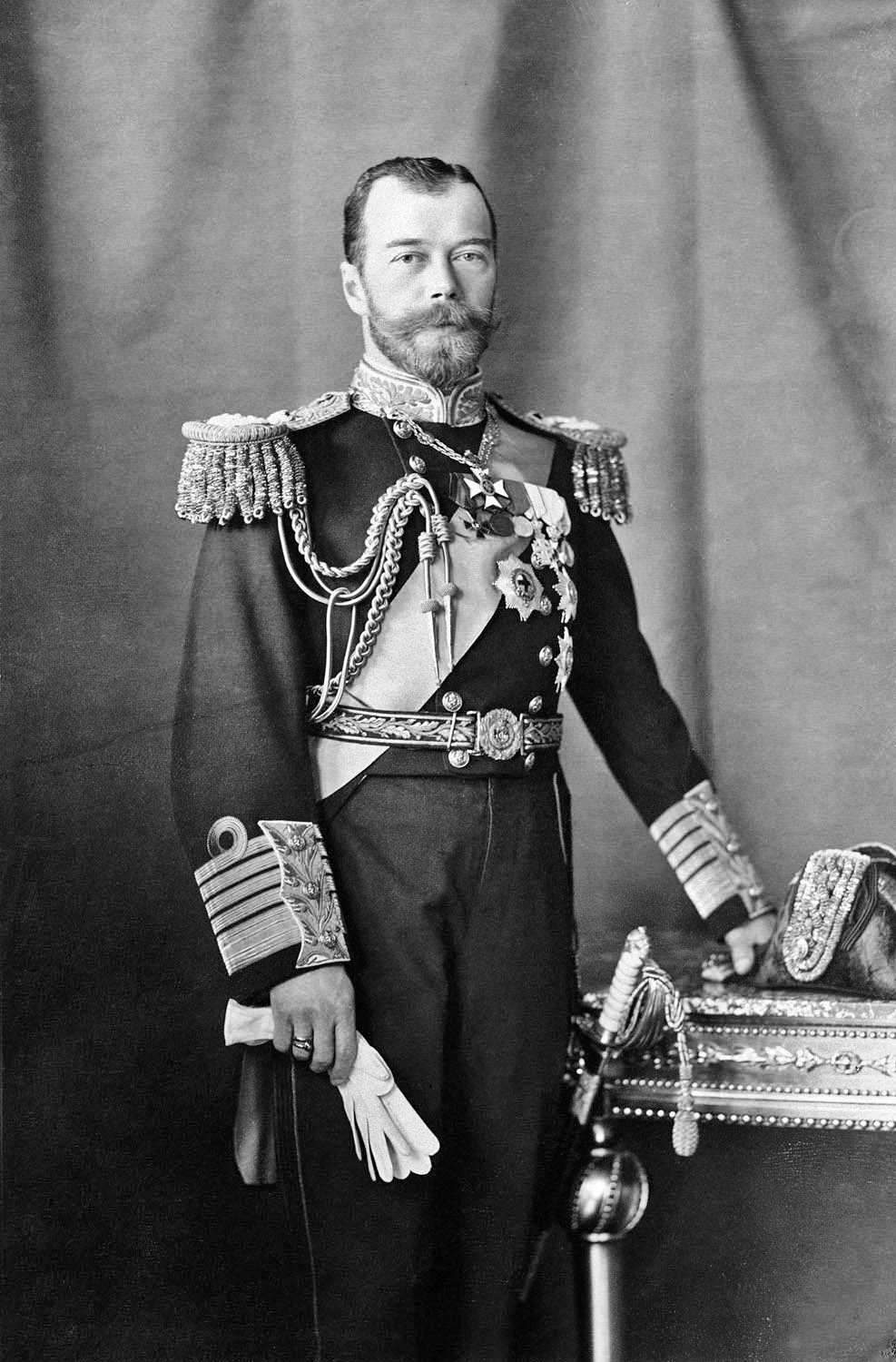 The tsar, a bearded man in richly decorated uniform, poses for the photography. He holds a pair of gloves in his right hand, the left hand rests on his hat on a side table.